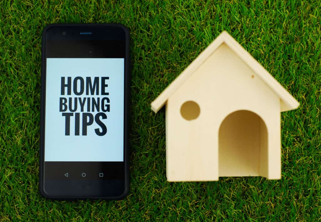 Home Buying Tips text on phone laying in grass next to wooden carving of a house.